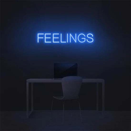 Blue neon sign displaying the word "FEELINGS" mounted on a dark wall above a desk with a chair and laptop Feelings neon sign