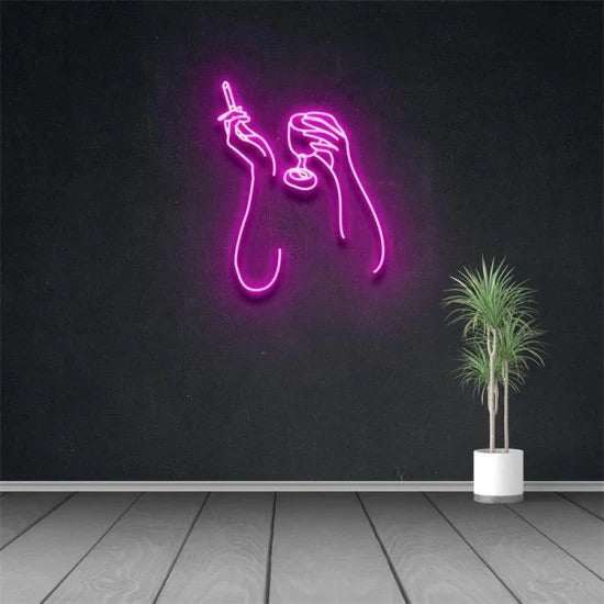 Pink neon sign of hands holding a cigarette and a drink on black wall above potted plant