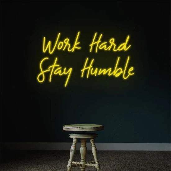 Yellow neon sign displaying "Work Hard Stay Humble" on dark wall above wooden stool