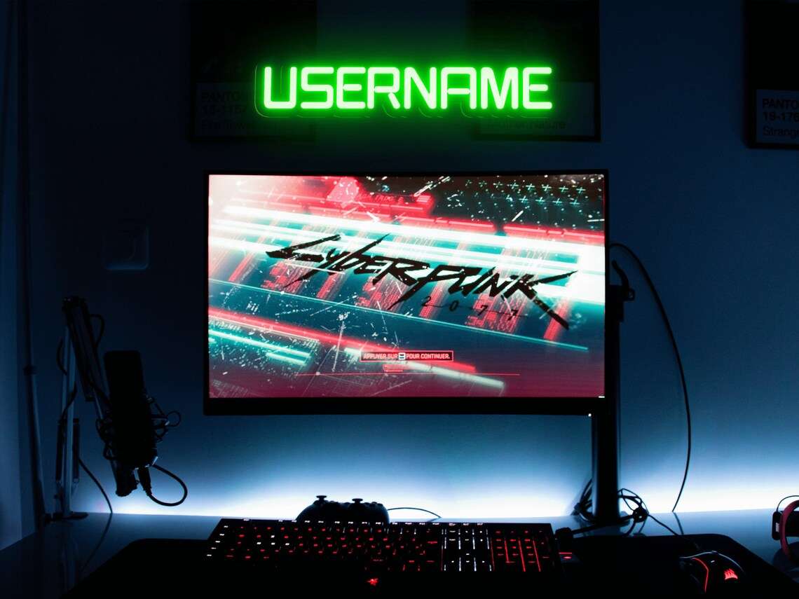 Green neon sign displaying the word USERNAME above a gaming setup with Cyberpunk 2077 on the monitor